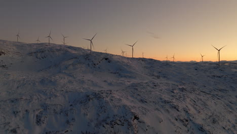 Drone-pan-shot-of-turning-wind-turbines-in-snowy-landscape-against-sunset-sky