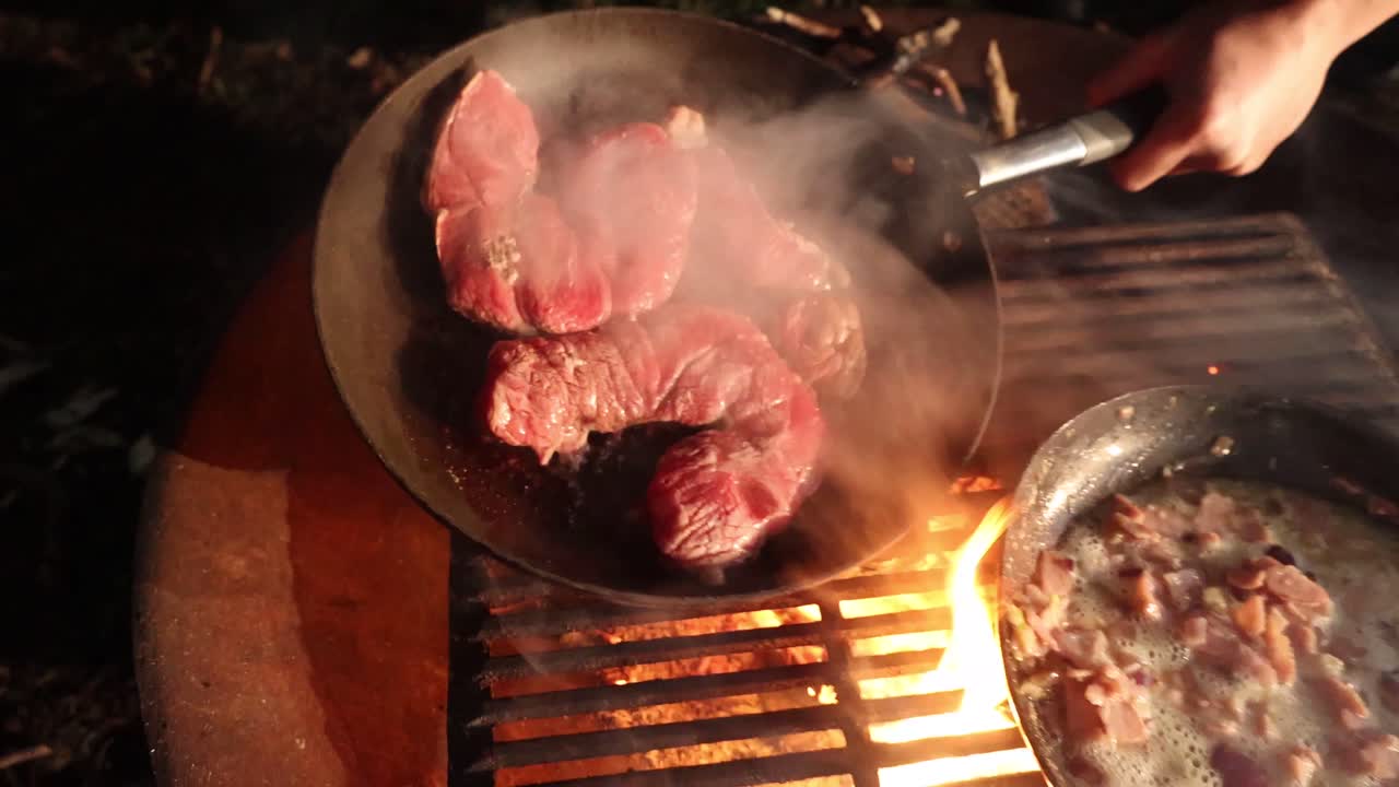 A close up of a big steak being grilled on a camp fire in the dark.