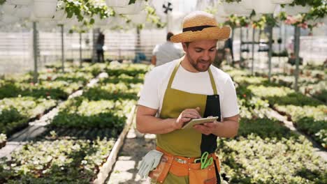 Hübscher-Männlicher-Gärtner-In-Einer-Schürze-Mit-Einem-Tablet-Geht-Um-Das-Gewächshaus-Herum-Und-Macht-Sich-Notizen