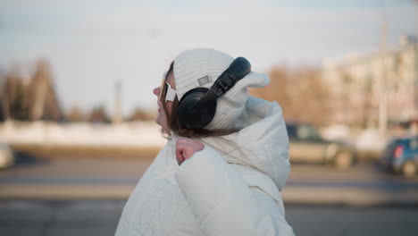 close up view of student spinning joyfully in winter park wearing white coat, beanie, headphones, and sunglasses, arm raised, smiling under sunlight with blurred cars passing in snowy background