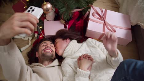 A-brunette-guy-and-his-girlfriend-in-a-white-sweater-lie-on-the-floor-and-take-a-selfie-near-the-New-Year-tree-and-pink-gifts-in-a-cozy-room-in-winter