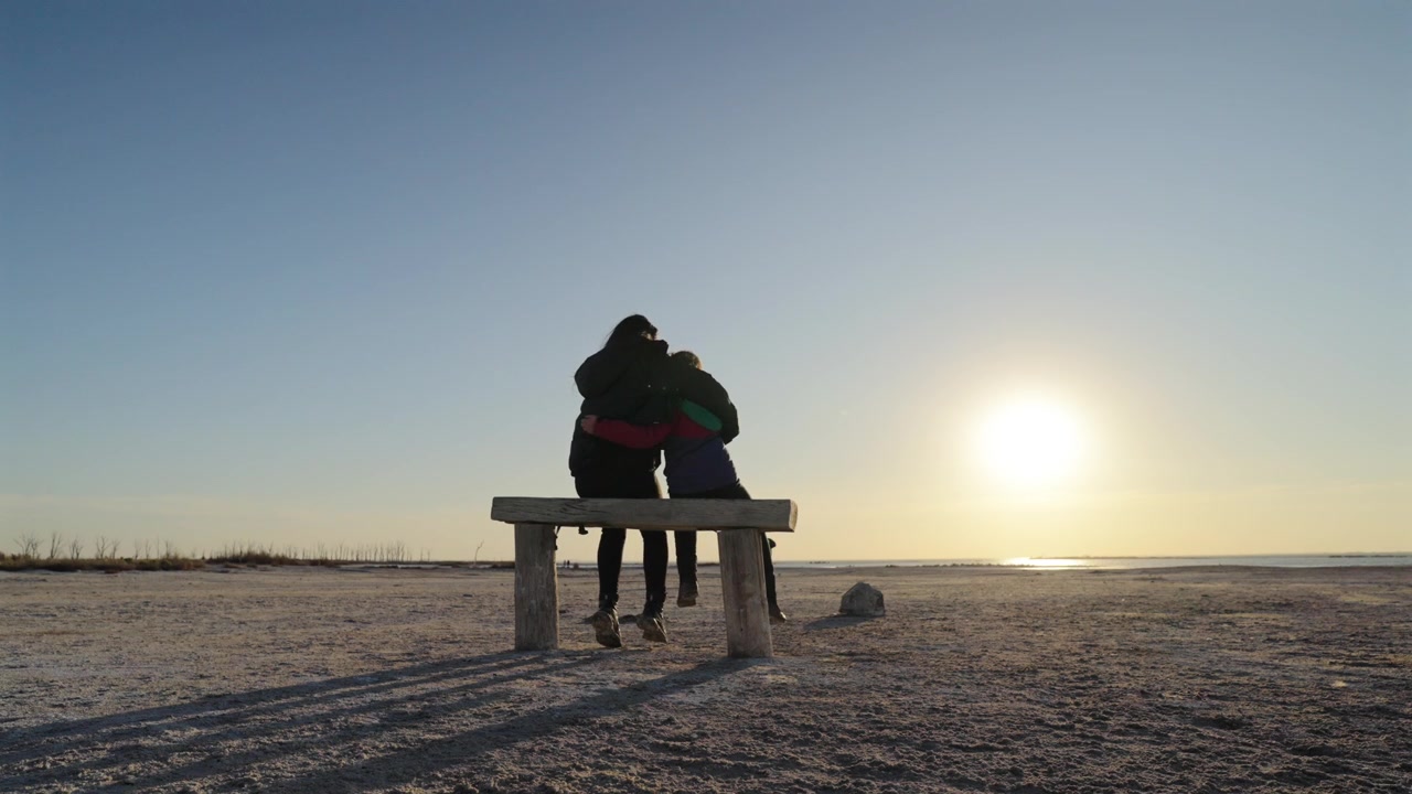 Two people meeting on bench at shoreline with one sitting and receiving an embrace under low sun creating long shadows and warm light suggesting support connection and affection static shot
