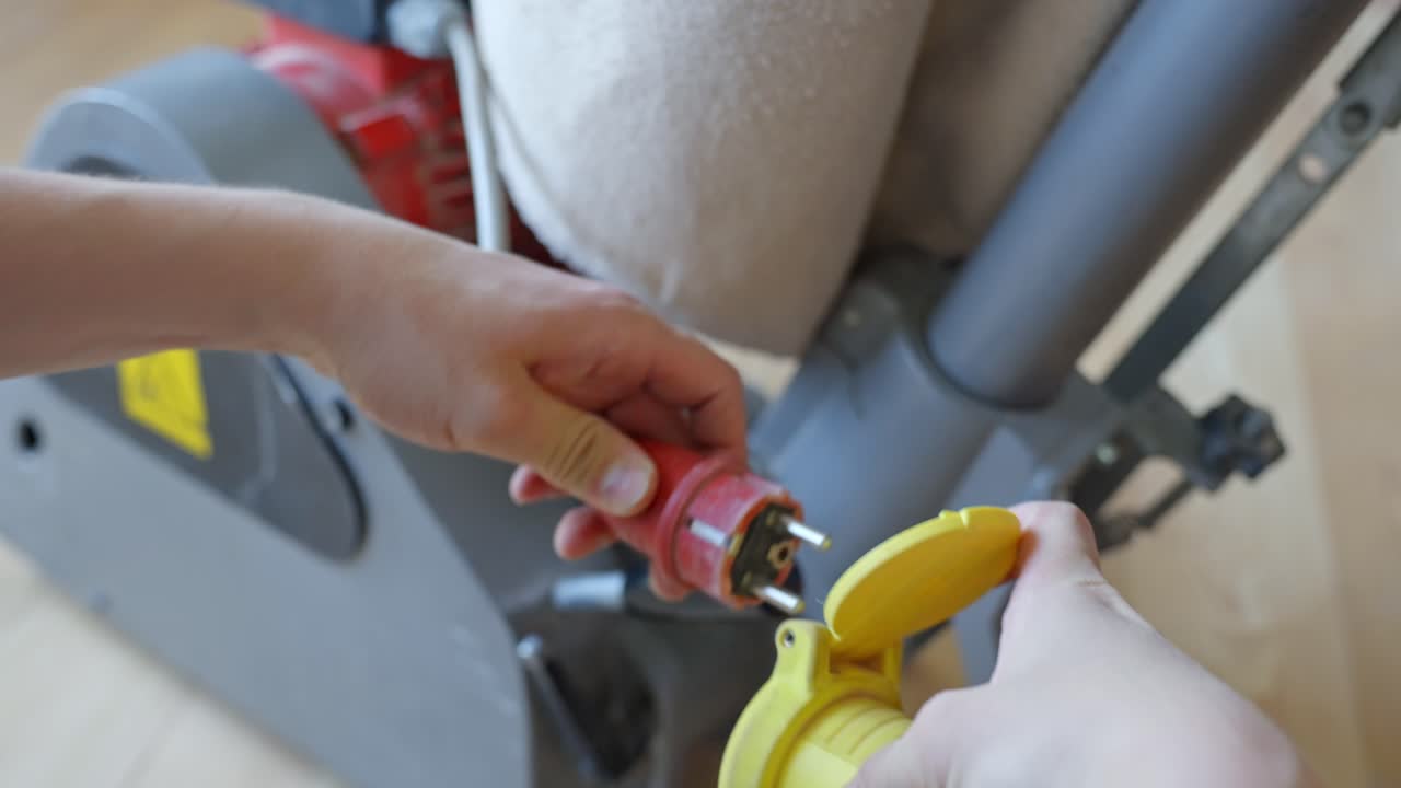 Hands prepare a wood floor sanding machine for operation by connecting its red industrial power plug to a yellow socket, readying equipment for sanding, static camera, real time, renovation context