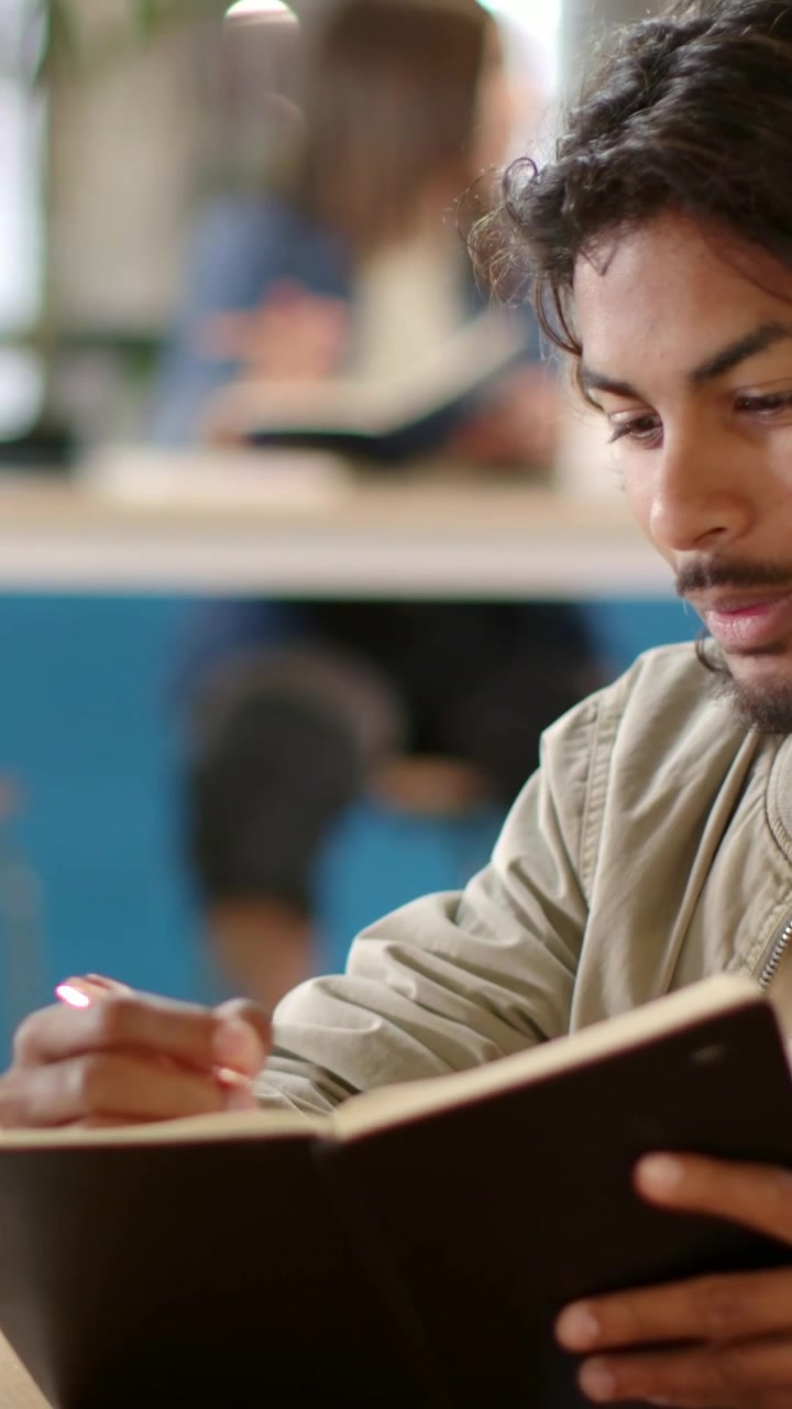 Video verticale: Studente che posiziona un quaderno, apre le pagine e prende appunti con una penna in un caffè del campus.