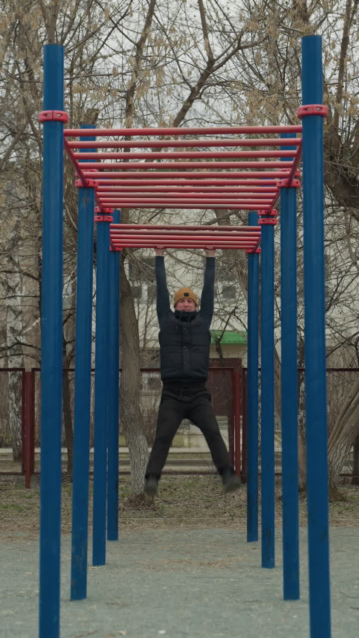 A coach jumps up to grab a red iron bar and begins swinging in an outdoor exercise area, in the background, a residential building, passing cars, and someone playing football can be seen