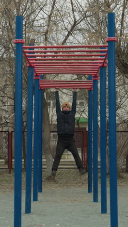 a coach jumps up to grab a red iron bar and begins swinging in an outdoor exercise area, in the background, a residential building, passing cars, and someone playing football can be seen