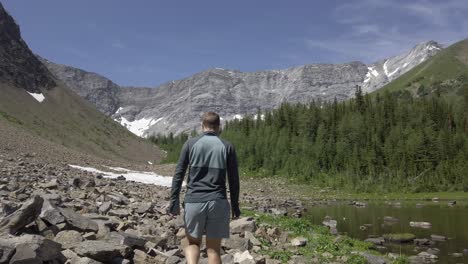 Hiker-walking-down-valley-by-pond-and-forest-followed-Rockies,-Kananaskis,-Alberta-Canada