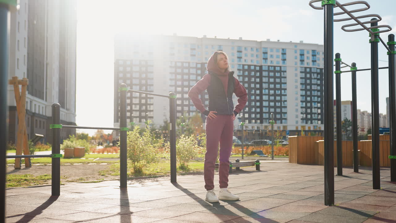 Female athlete stands with hands on waist in sunny urban park by bar, rolling neck slowly to warm up muscles under clear sky before starting dynamic outdoor workout routine