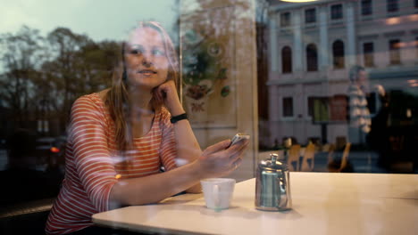 Mujer-Con-Teléfono-En-La-Cafetería-Disfrutando-De-La-Vista-Exterior.