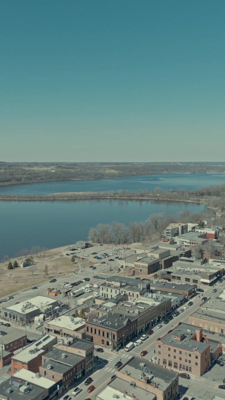 Ripresa aerea con parallasse verticale sopra il centro di Hudson, Wisconsin, che ruota per rivelare la storica griglia di Main Street, il parco sul lungomare e l'ampio fiume St. Croix sotto un cielo primaverile limpido.