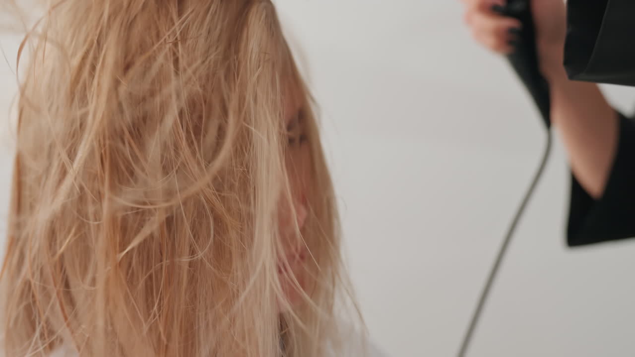 Sesión de secado en salón. Cliente caucásica con el pelo mojado y encrespado. El estilista seca el pelo con un secador de alta velocidad. Las hebras se levantan con el flujo de aire, creando volumen texturizado. Primer plano en un estudio luminoso.