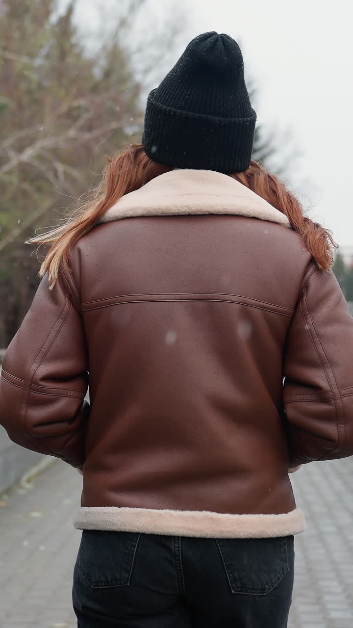 Back shot of student in black cap brown jacket walking slowly with hands in pockets on wide stone pathway through urban park surrounded by pine trees modern buildings and cloudy sky