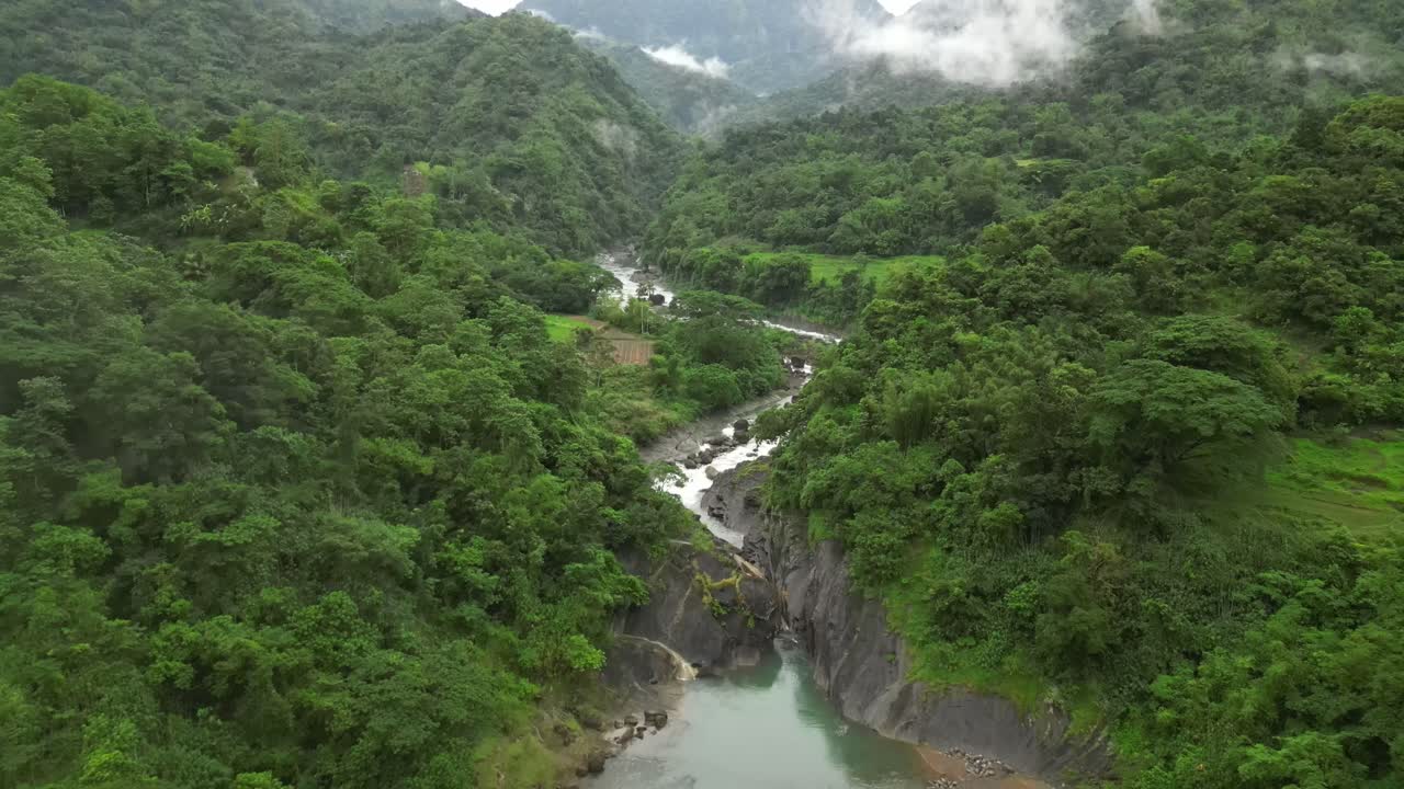 A wide aerial angle drifts to a final hold over mist-covered peaks and a winding river. Dense forest blankets the valley as rocky terrain and steep slopes settle into the layered landscape