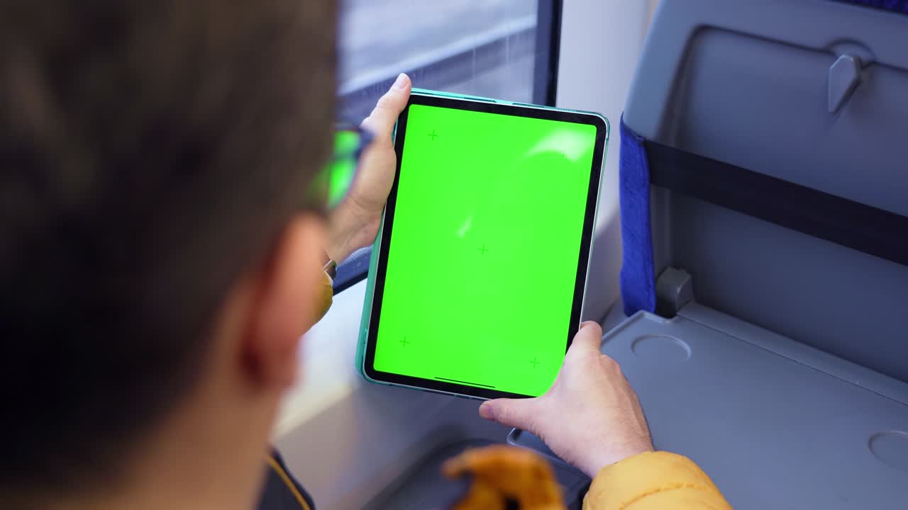 Mand set bagfra, der ser på en tablet-pc med grøn skærm og browser online i et tog. Mockup til visning af indhold på en tablet. Tom digital telefon i hånden på en fyr, der sidder indendørs i offentlig transport.
