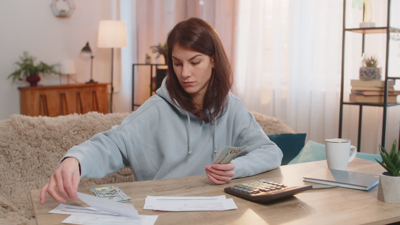 Young woman counting money while sitting on sofa with receipts and calculator on tablet at home