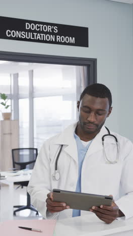 vertical video of happy african american male doctor using tablet in hospital, slow motion