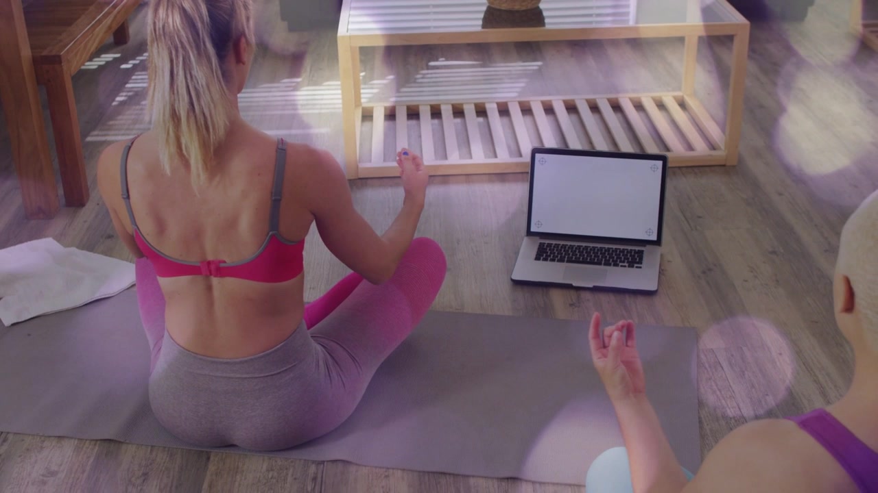 Mulher entrando em meditação em um tapete cinza enquanto segue uma sessão de bem-estar no laptop, com o parceiro espelhando