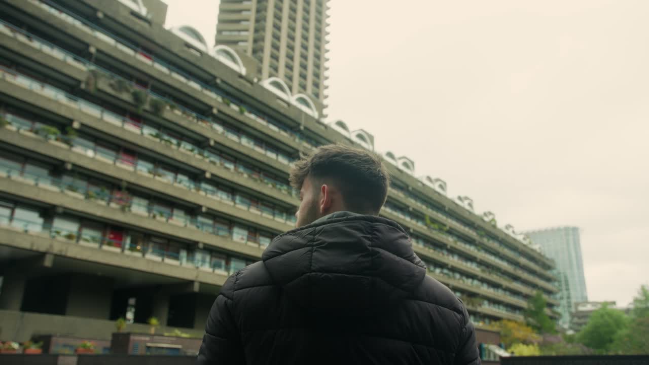 Medium rearview of man figure in black puffer jacket under concrete ledges in Barbican Estate, London housing architecture