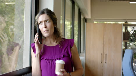 caucasian casual businesswoman drinking coffee and talking on smartphone in office, slow motion