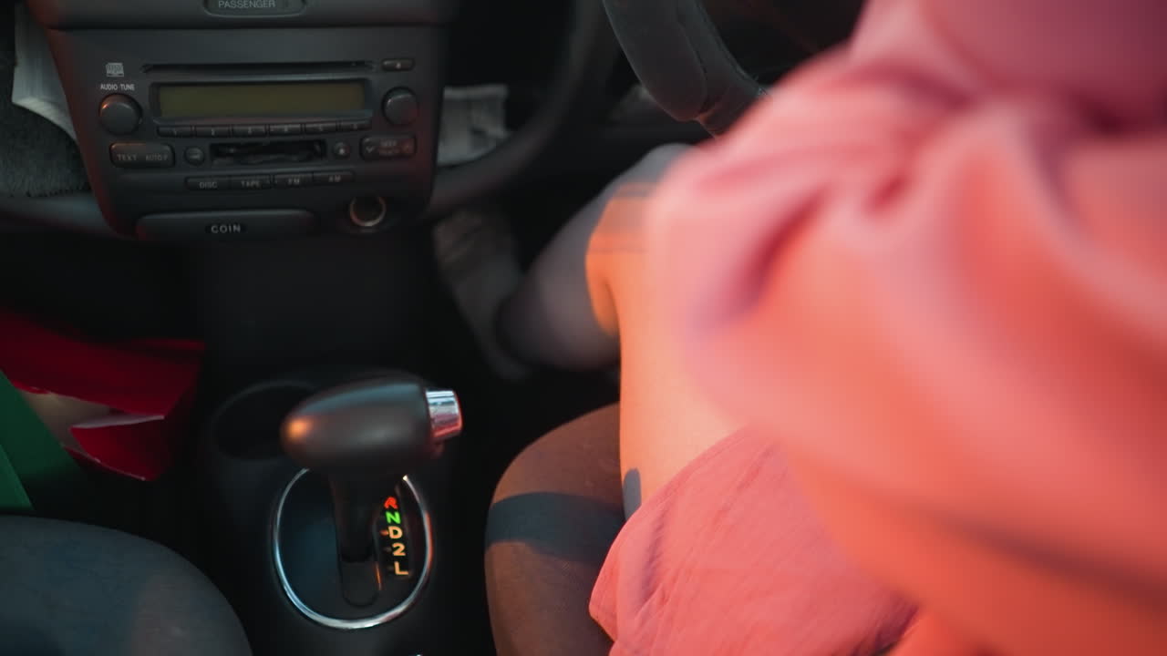 женщина в розовом платье, сидящая в своей машине, хватает чашку кофе возле переключения передач и глотает, интерьер машины слегка размыт мягкими отражениями проходящих огней, освещающих ее.