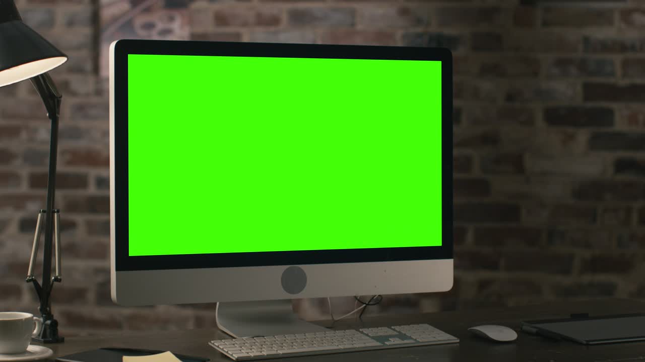 imágenes de un monitor de computadora con pantalla verde de pie en una mesa junto a una tableta, lámpara, taza de café, portátil y ratón en un loft.