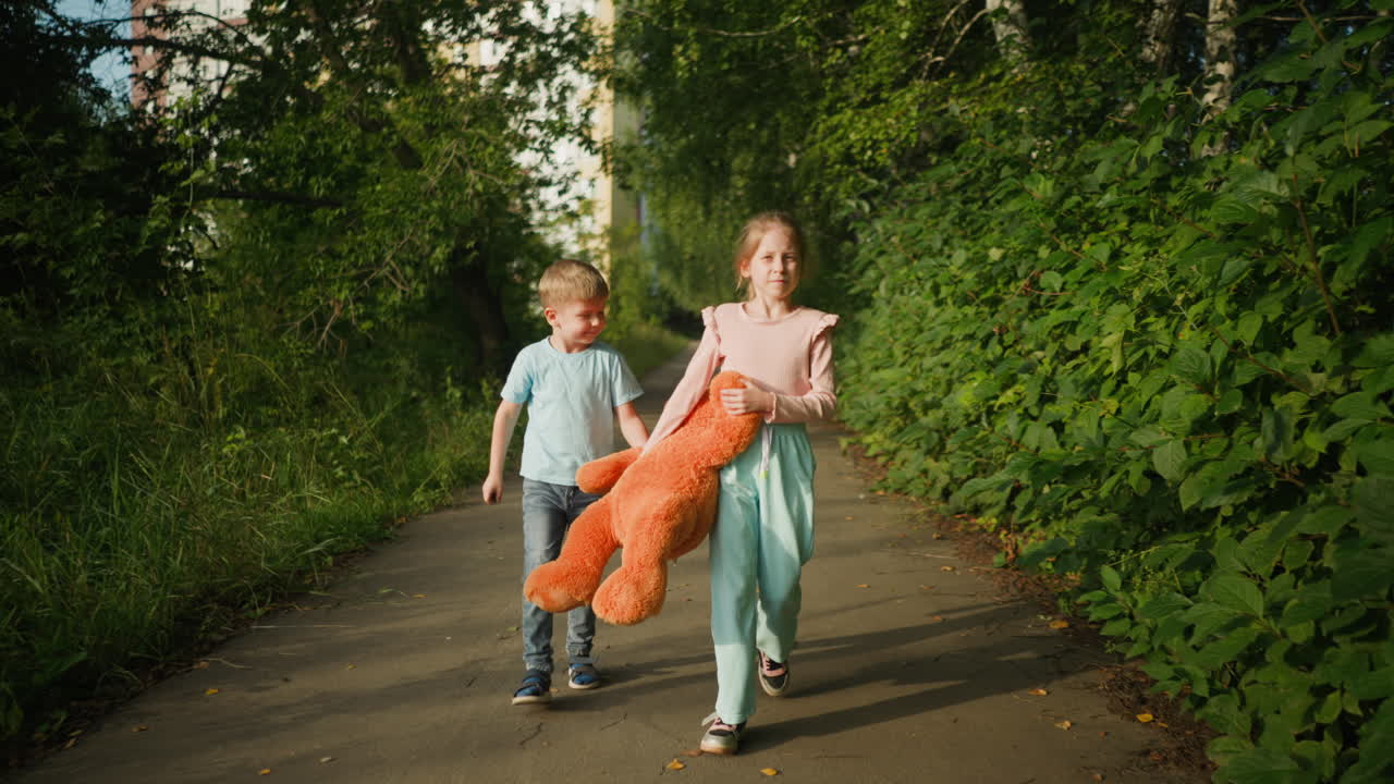 Two kids walking side by side on paved path bordered by greenery, holding large orange teddy bear off ground, with soft afternoon sunlight casting distinct shadows of children and toy on ground