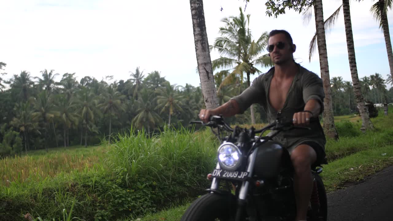 Young, fit and cool man is driving on the countryside roads of Bali with his custom motorcycle with beautiful rice fields and local people surrounding him