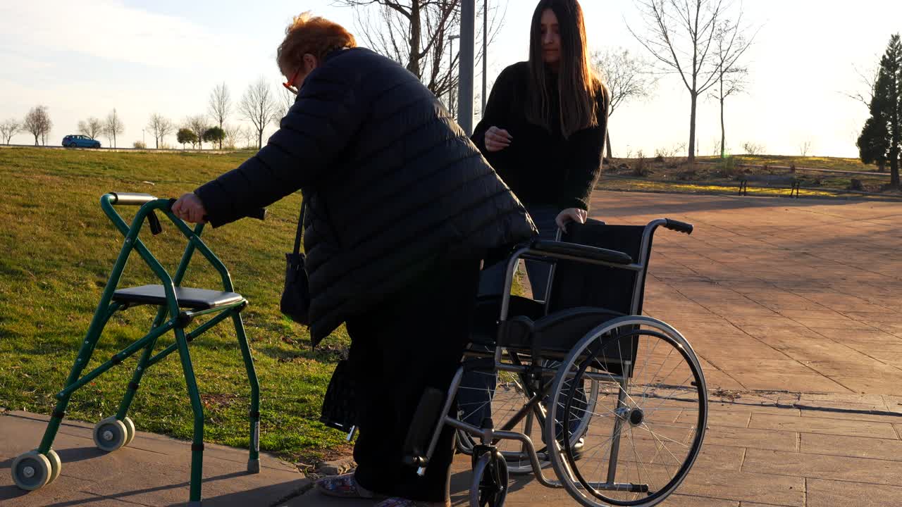 Elderly woman switching from walker to wheelchair with help outdoors