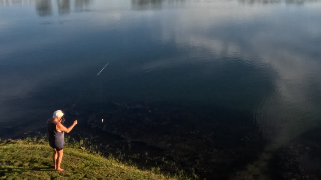 A person casts a fishing line beside a reflective lake, surrounded by lush greenery.
