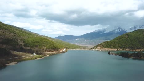 drone shot over a forest and a lake with a snowed mountain peak in the background, greece