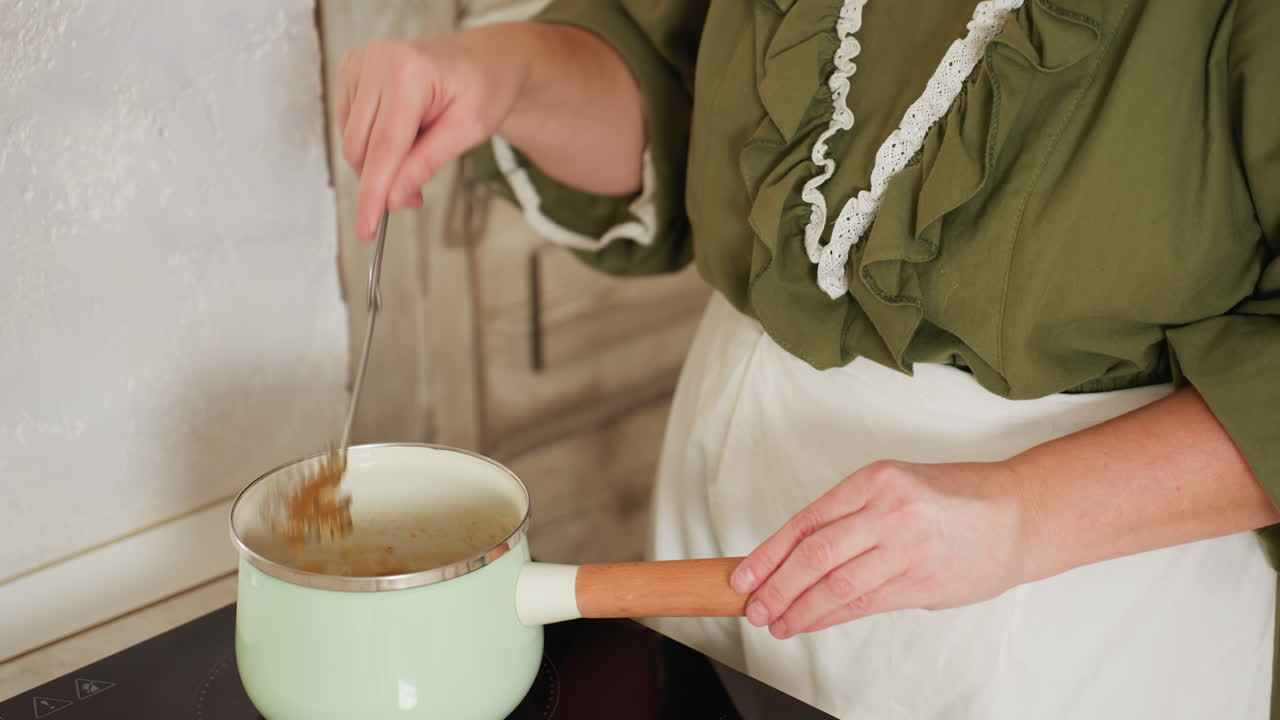 Close-up partial view of chef in green ruffled dress and white apron preparing soup, holding ladle dripping liquid into pot in rustic kitchen with wooden door in background
