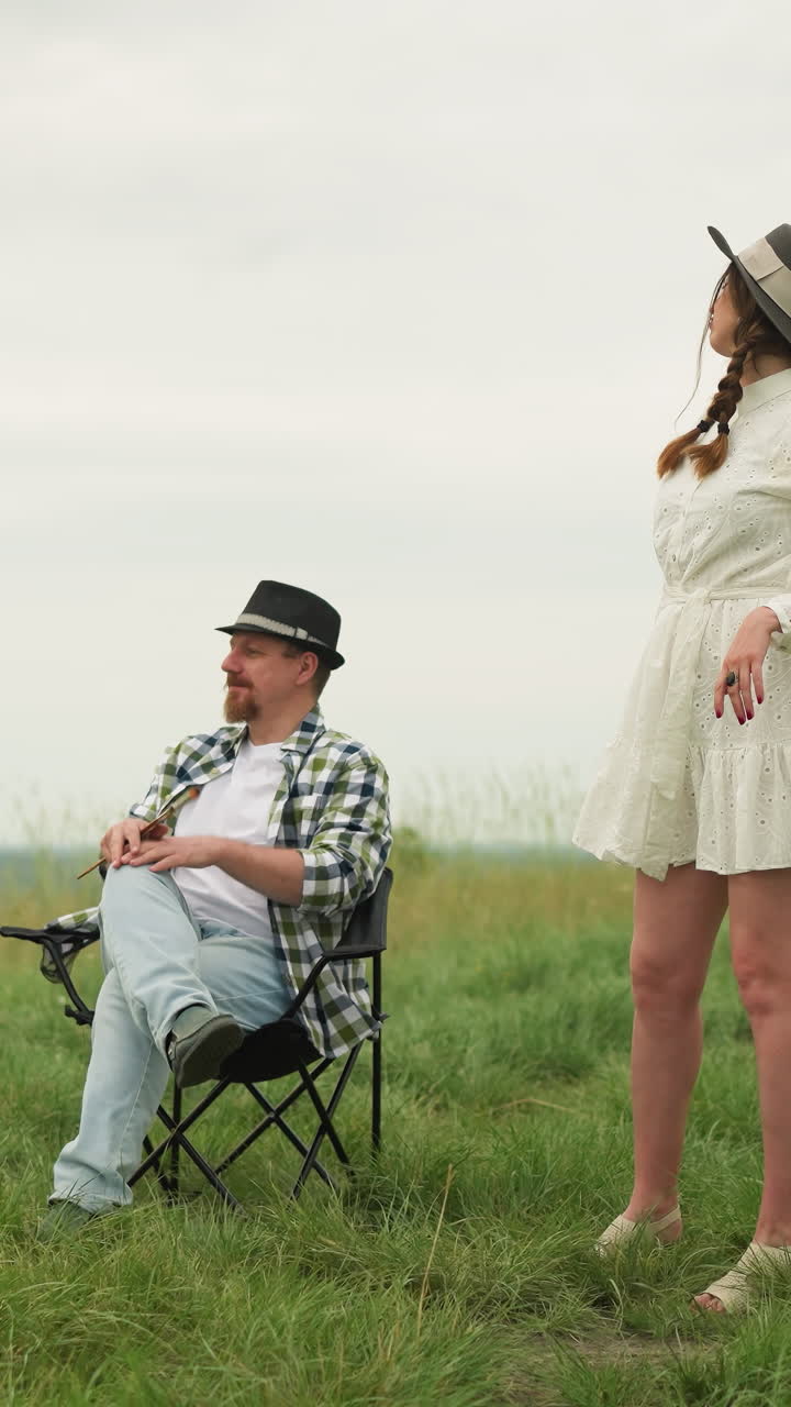 색 셔츠를 입은 한 예술가가 잔디에 있는 의자에 앉아 을 들고 다리를 고 색 드레스를 입은 여자에게 앉아있는 자세를 보여주고 있다.