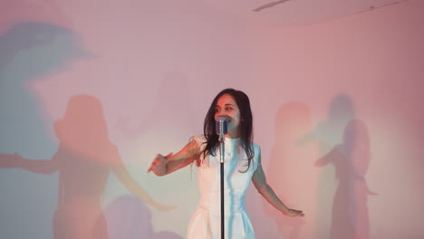 vocalist in a white gown singing passionately, lifting hands up as the camera zooms in against a colorful background with dynamic lighting and shadows