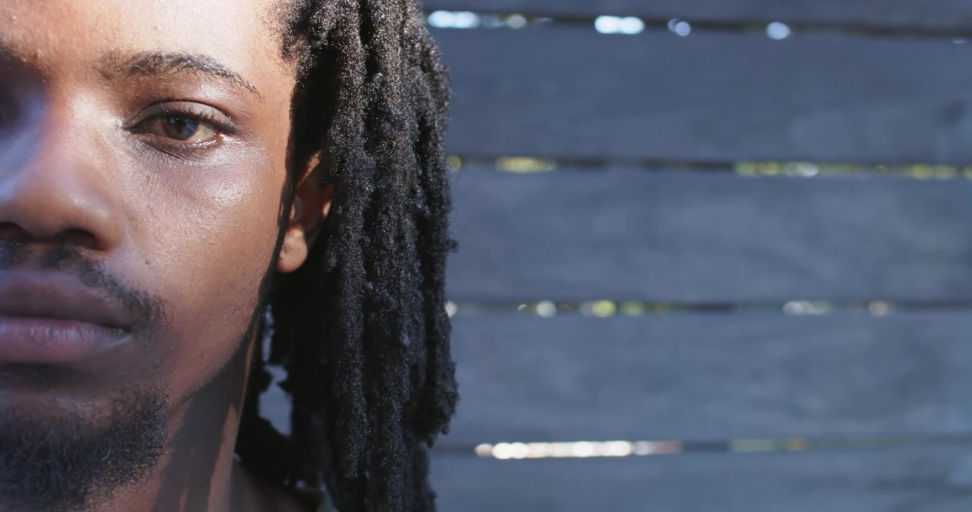 Uomo africano con i dreadlock che sbatte le palpebre e sposta lo sguardo verso una recinzione a doghe dipinta dalla luce del sole, con spazio per il testo.