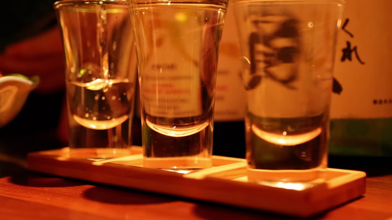 Close-up of sake being poured into two glasses on a wooden tray in warm lighting.