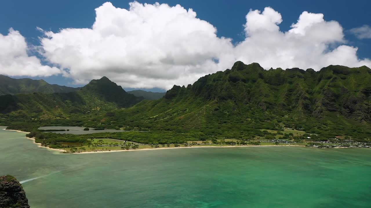 Aerial drone view showing Kauai coastline with lush green mountains, sandy beach, and clear turquoise ocean water under partly cloudy sky, capturing the island’s natural tropical beauty