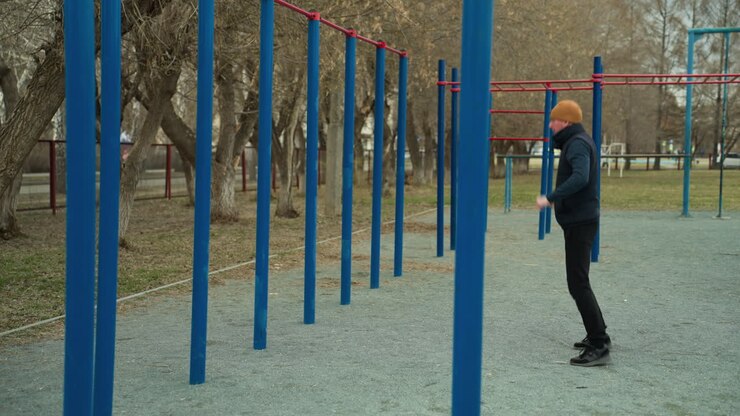 A coach exercise alone in a stadium, wearing a brown beanie and black outfit, he's surrounded by blue and red iron bars, with trees and distant cars and people passing by