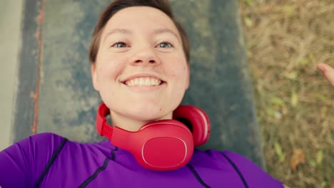 First-person-view:-A-happy-girl-with-a-short-haircut-with-brown-eyes-in-a-purple-top-and-red-headphones-lies-down-on-a-bench-and-takes-a-selfie-in-a-skatepark-in-summer