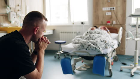 mid-aged caucasian man sits in the hospital ward. pregnant woman lies in bed at backdrop in blur. husband worries about his wife before labor.
