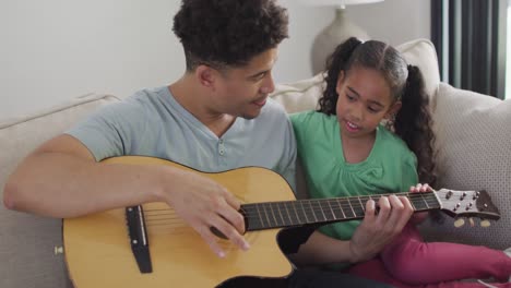 padre y hija biraciales felices sentados en el sofá tocando la guitarra