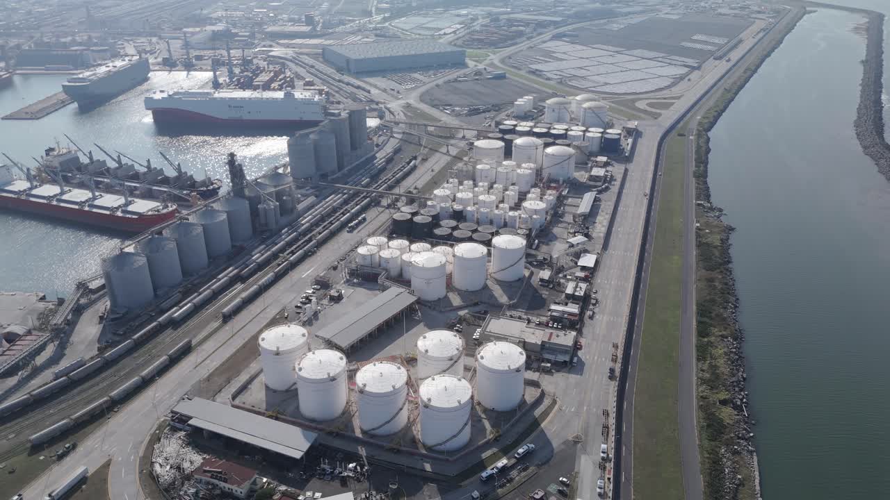 Wide aerial view of a major port facility featuring docked cargo ships, extensive oil storage tanks, grain silos, and rail connections. Clear weather enhances visibility of the industrial layout.