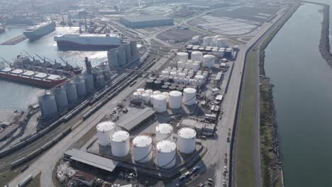 wide aerial view of a major port facility featuring docked cargo ships, extensive oil storage tanks, grain silos, and rail connections. clear weather enhances visibility of the industrial layout.