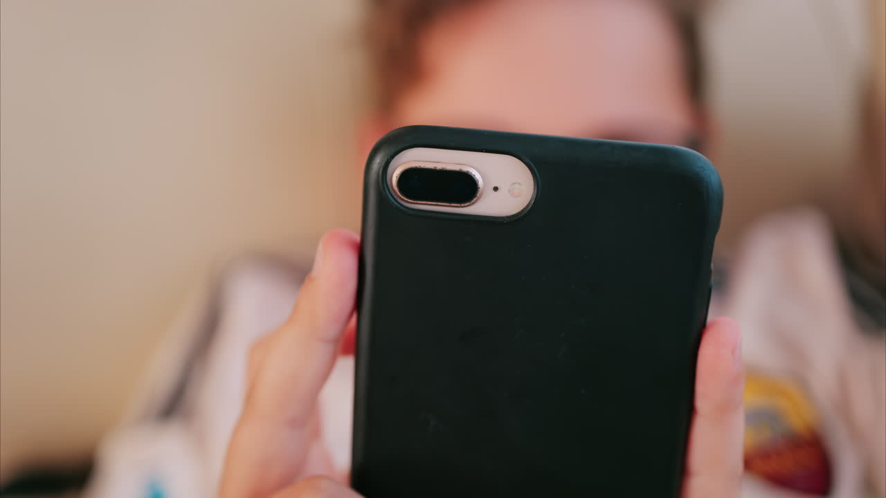 Close up of a child holding a smartphone with a black case while lying down indoors