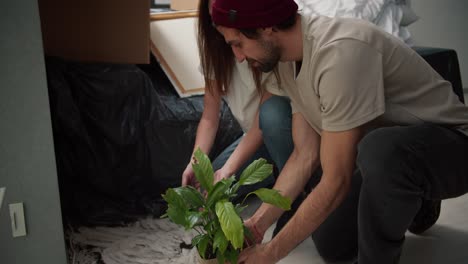 un hombre moreno con paja en una camiseta beige junto con su novia morena en una camisa blanca recogen tierra de una flor de la casa que cayó en el suelo después de mudarse a un nuevo apartamento entre cajas y muebles en polietileno negro en un nuevo apartamento