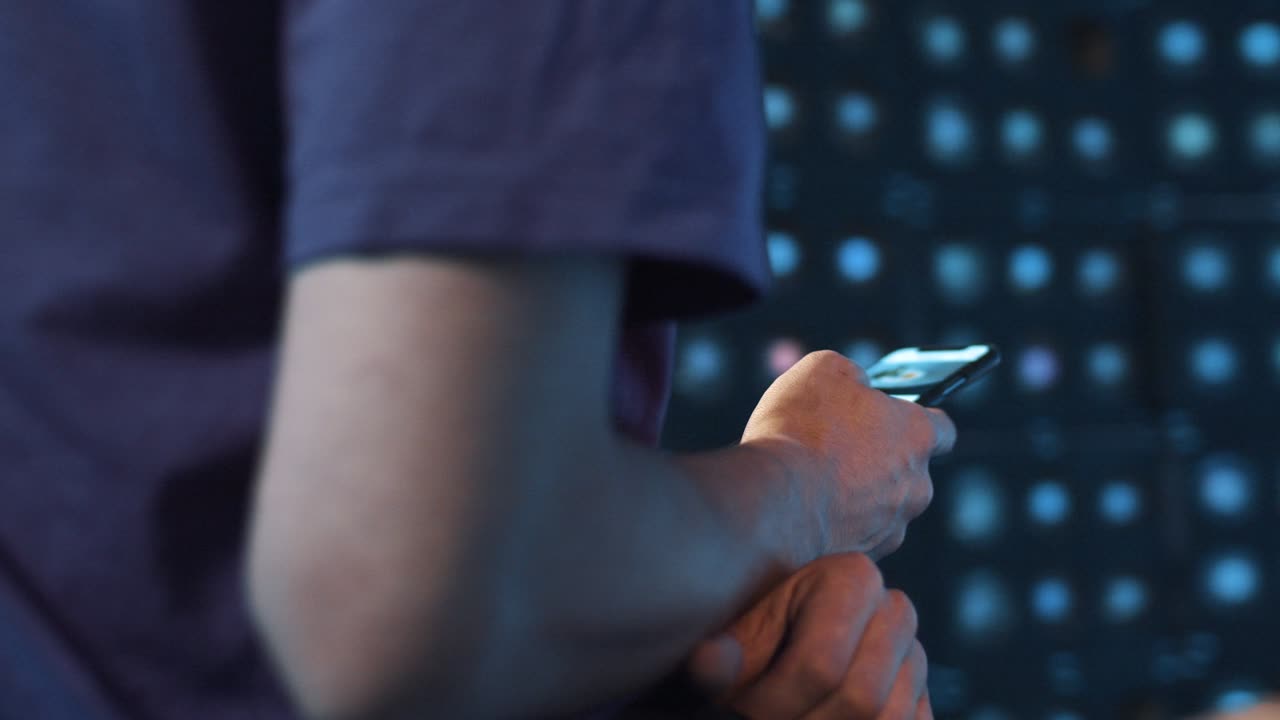 Slim male in violet t-shirt sits in dark room and actively using his smartphone.