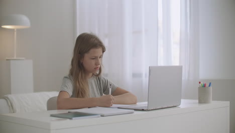 school girl is learning online at home viewing at display of notebook and writing at copybook e-learning and homeschooling at pandemic of coronavirus