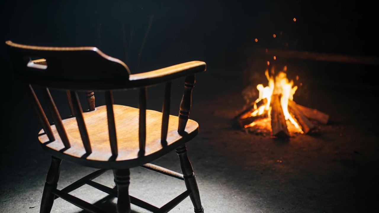 Flickering fire from logs lighting wooden spindle-back armchair on cabin floor sparks rising