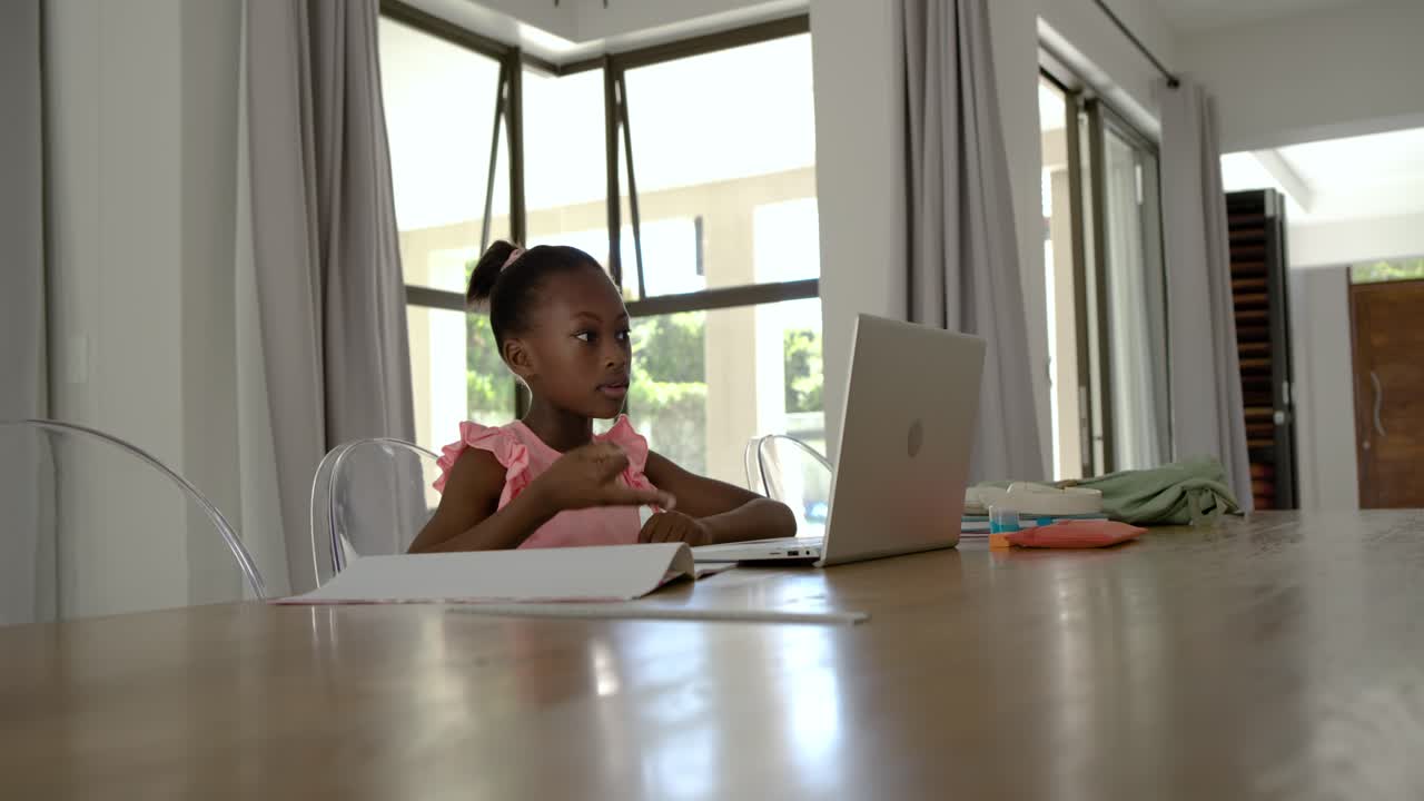 African child in pink top resting chin on pencil, writing notes, checking laptop at home, studying