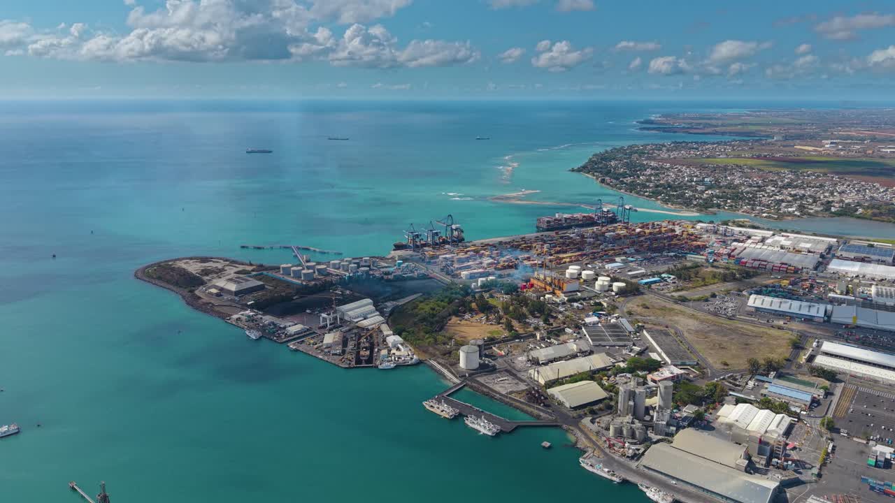 Aerial of the Port Louis industrial harbor and container terminal in Mauritius. The shot captures a busy commercial port with cranes, cargo ships, and containers by the turquoise ocean
