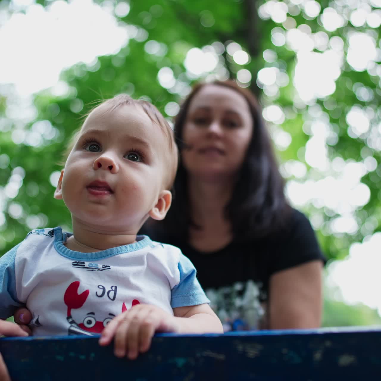 Healthy Caucasian child spending time outdoors. Cute kid standing on the bench looking curiously around. Close up. Mom at backdrop. Low angle view